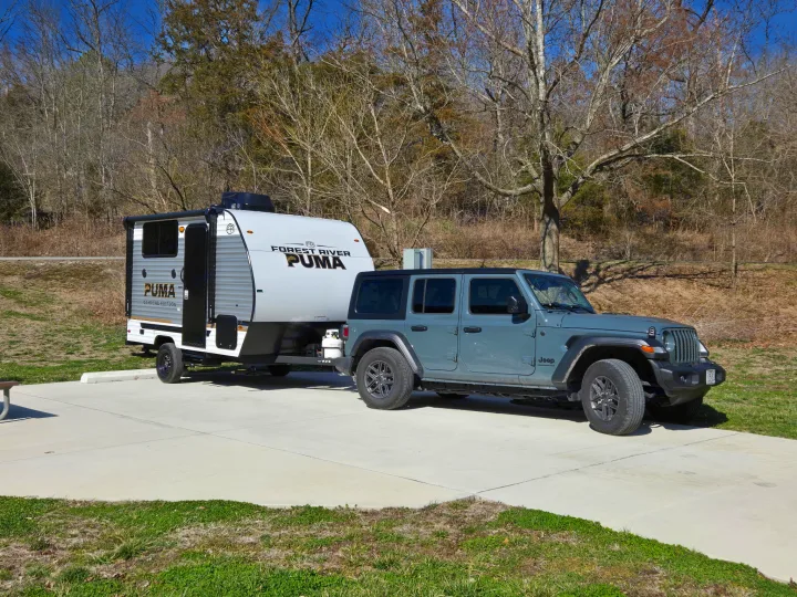RV rental: A gray Jeep Wrangler tows a Forest River Puma RV on a paved campsite, ready for adventure among the trees.. Palomino Puma Camping Edition 122BHCE 2026