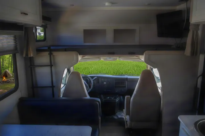 RV rental interior featuring a front cab, overhead bunk, dinette, and a window view of a campsite and vibrant green field.. Gulf Stream Conquest 2024