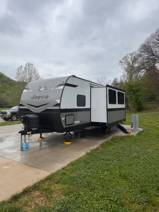 A grey Jayco RV rental travel trailer is parked with stabilizers and an extended slide-out at a paved campsite on a cloudy day.. Jayco Jay Flight SLX 2024