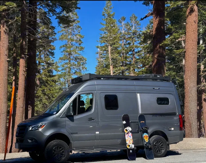 Gray Mercedes Sprinter RV rental camper van with snowboards & skis, parked among tall pines for winter outdoor adventure.. Winnebago Revel 2025