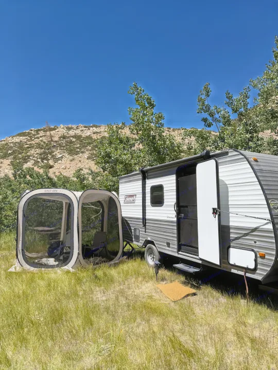 RV rental travel trailer parked in a sunny, grassy campsite next to an EAST OAK screen tent, with mountains behind.. Coleman 17B 2024