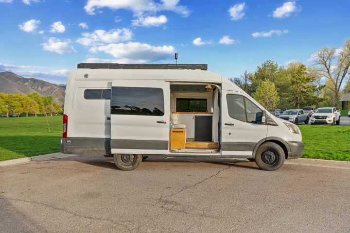 White campervan RV rental with open door revealing interior, parked in front of mountains & a green field under a blue sky.. Ford Van 2015