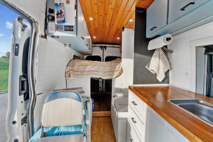 This custom-built RV rental interior features a compact kitchen, comfortable bed, wood paneling, and an open side door.. Ford Van 2015