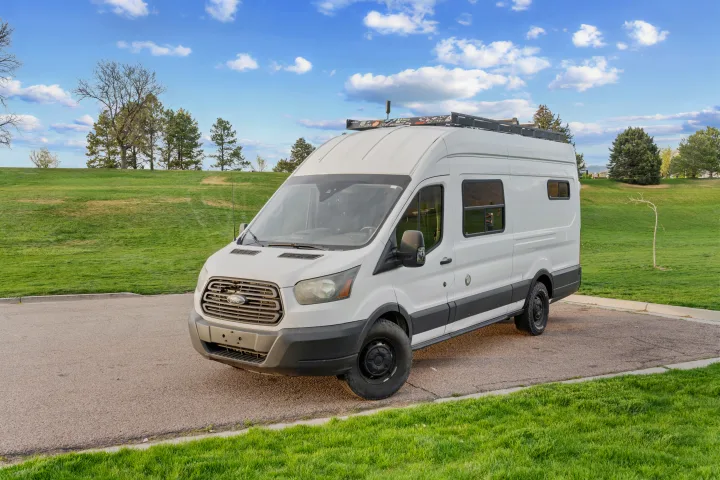 Explore in this white Ford Transit camper van, an ideal rv rental. It's parked on asphalt next to a green hill under a blue sky.. Ford Van 2015