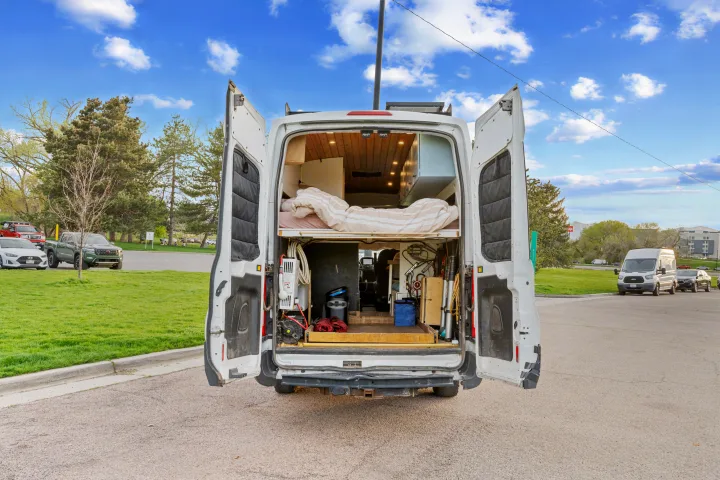 White camper van's rear doors open, showcasing a cozy bed, organized storage, and utilities inside, perfect for RV rental adventures.. Ford Van 2015