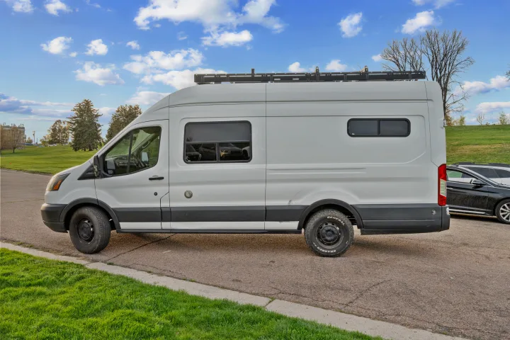 White Ford Transit campervan with custom windows and roof rack, available for RV rental. Perfect for your next adventure.. Ford Van 2015