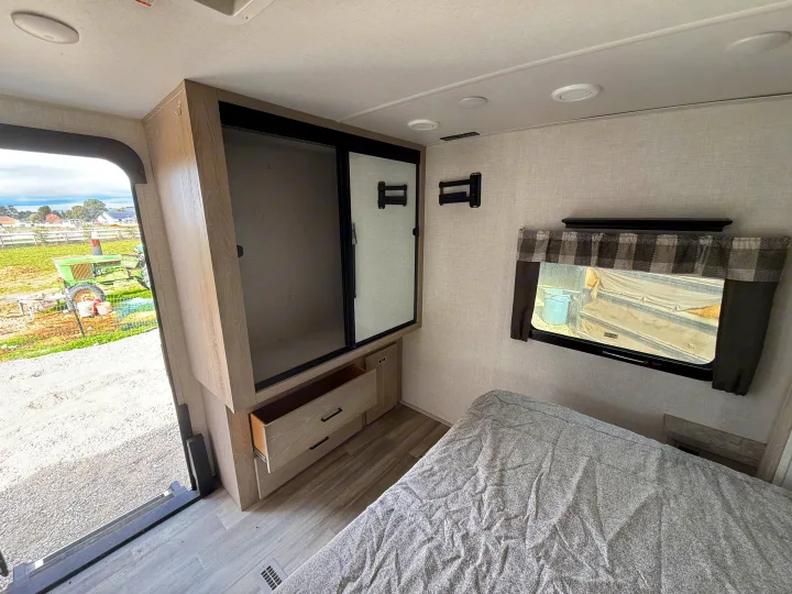 RV rental bedroom: a cozy bed, open wardrobe with drawers, and a window framing a rural view with a green tractor.. Forest River Flagstaff Super Lite 2021