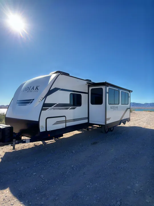 White Kodiak Ultra Lite rv rental parked on gravel, sun shining, with a lake and mountains in the background.. Dutchmen Kodiak Ultra Lite 2021