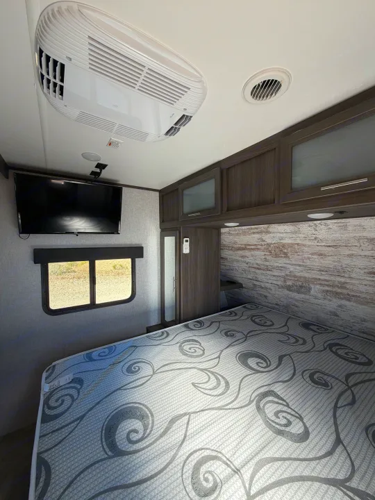 RV rental bedroom with patterned mattress, RV AC unit, TV, and wood-look cabinets. Cozy, modern travel trailer interior.. Dutchmen Kodiak Ultra Lite 2021