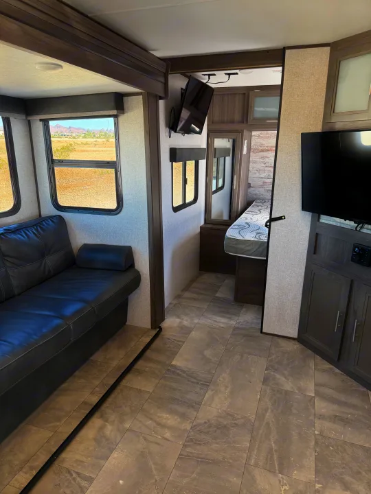 Modern rv rental interior with a dark leather sofa, TV, and bedroom, offering scenic desert views through large windows.. Dutchmen Kodiak Ultra Lite 2021