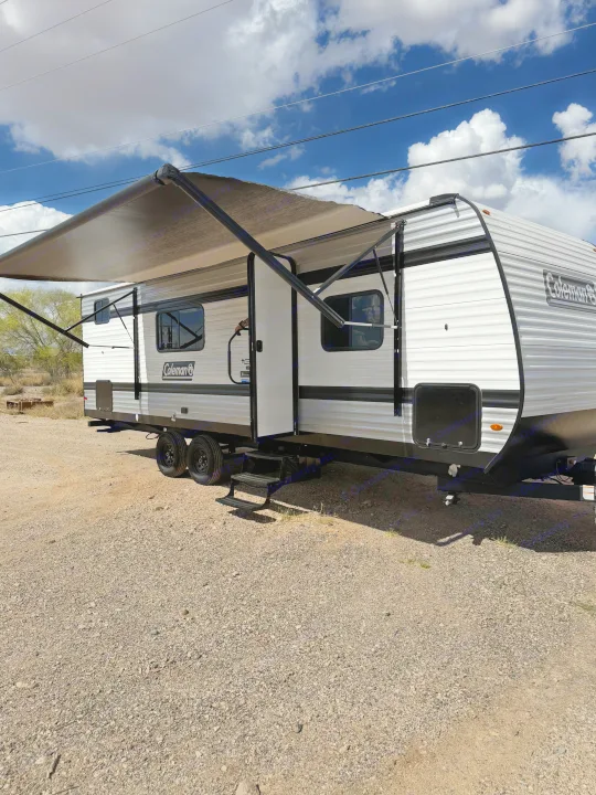 Coleman travel trailer with extended awning, available for rv rental, parked on gravel under a cloudy sky.. Keystone Coleman 2026