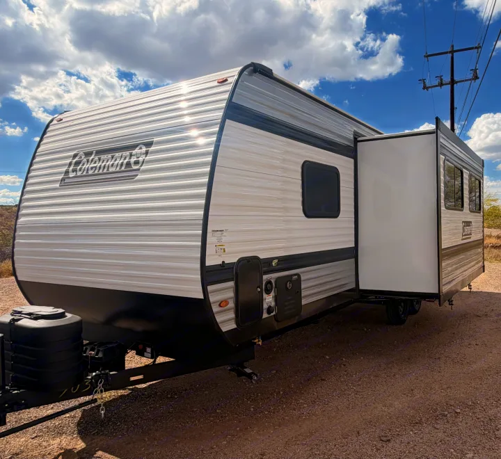 Side view of a Coleman rv rental travel trailer with its slide-out extended, parked on gravel under a cloudy sky.. Keystone Coleman 2026
