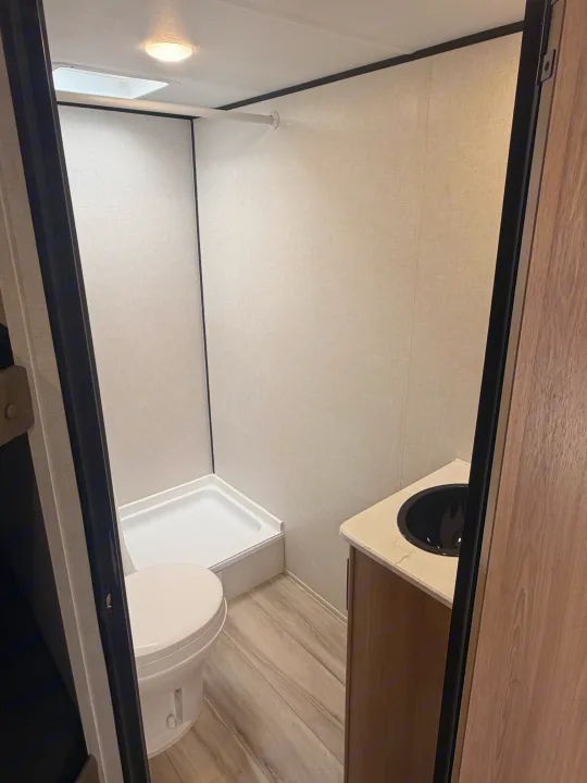 Compact rv rental bathroom features a small shower, toilet, & vanity with black sink. Light walls & wood-look floor.. Keystone Coleman 2026