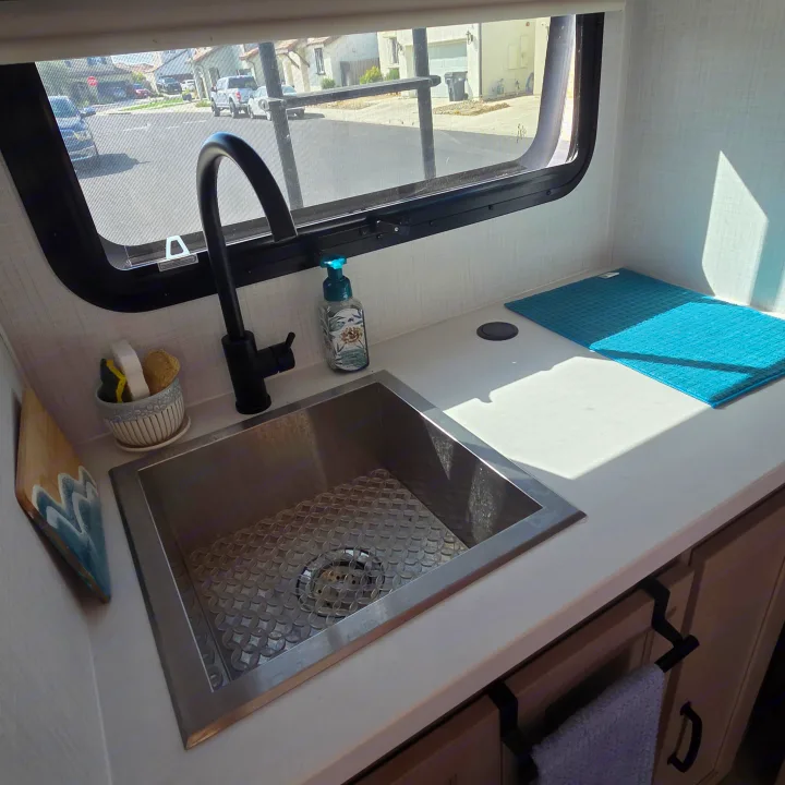Bright rv rental kitchen sink with black faucet, soap, sponges, a blue drying mat, and a window view of a residential street.. Forest River Rockwood Geo Pro 2024