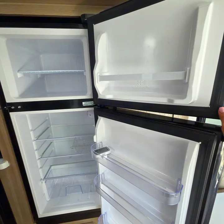 Inside an RV rental: empty, open refrigerator and freezer unit showing shelves & a thermometer.. Forest River Rockwood Geo Pro 2024