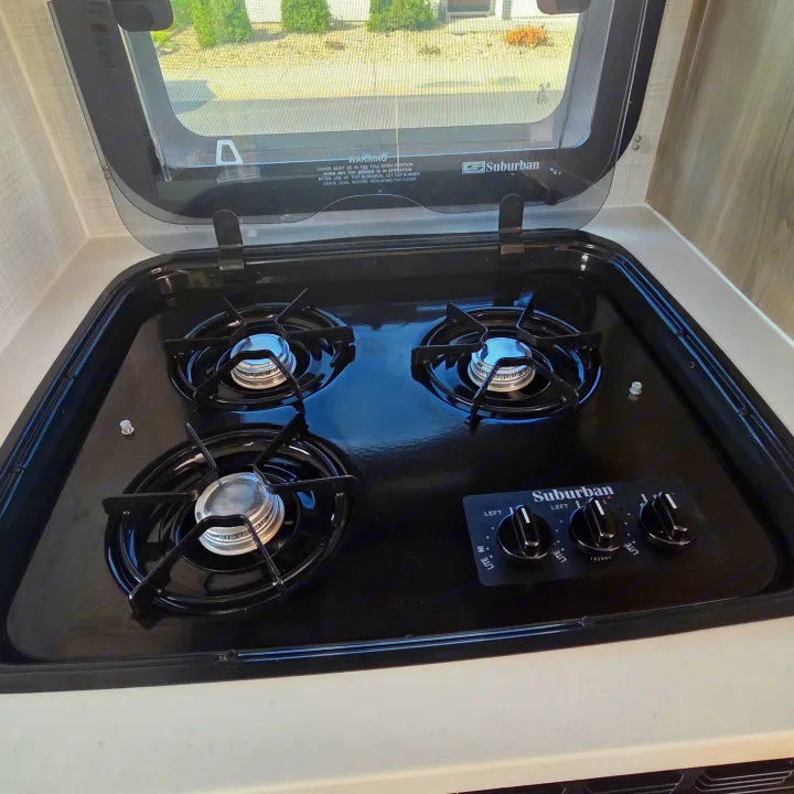 RV rental kitchen with a Suburban 3-burner gas cooktop, controls, and open glass cover. Ready for cooking on the go.. Forest River Rockwood Geo Pro 2024