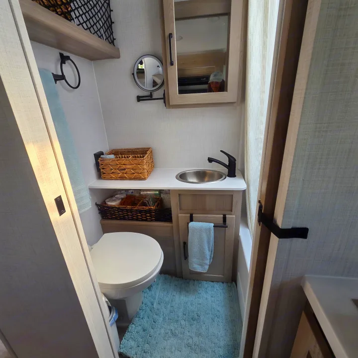 Compact RV rental bathroom with a toilet, sink, mirrored cabinet, and small magnifying mirror. Light wood cabinetry and storage baskets are visible.. Forest River Rockwood Geo Pro 2024