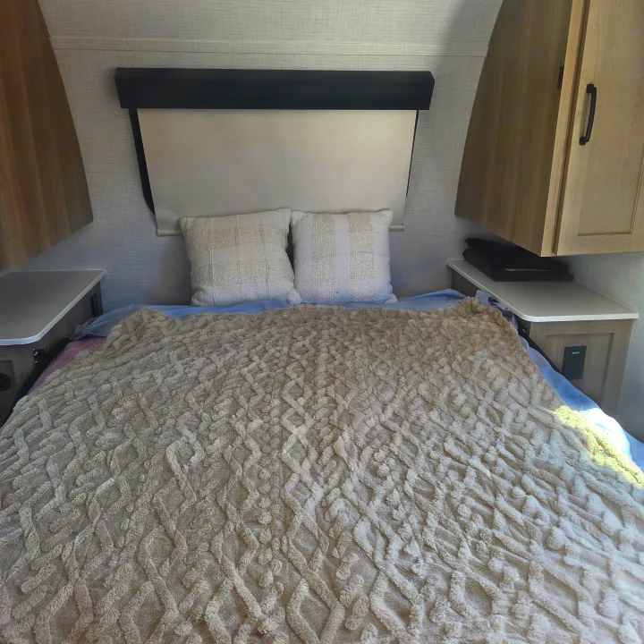 An RV rental bedroom with a comfortable bed, a textured beige blanket, two decorative pillows, and light wood cabinetry.. Forest River Rockwood Geo Pro 2024