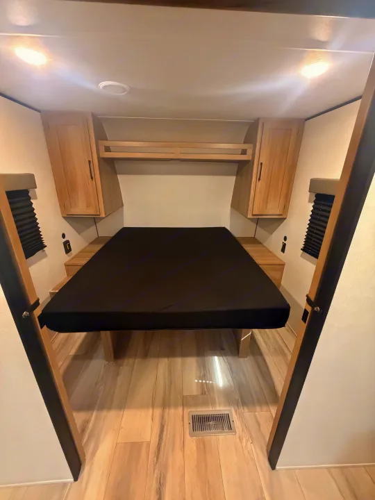 Inside an RV rental: a compact bedroom with a black mattress, light wood cabinets, and bright overhead lights.. Keystone Coleman 2026