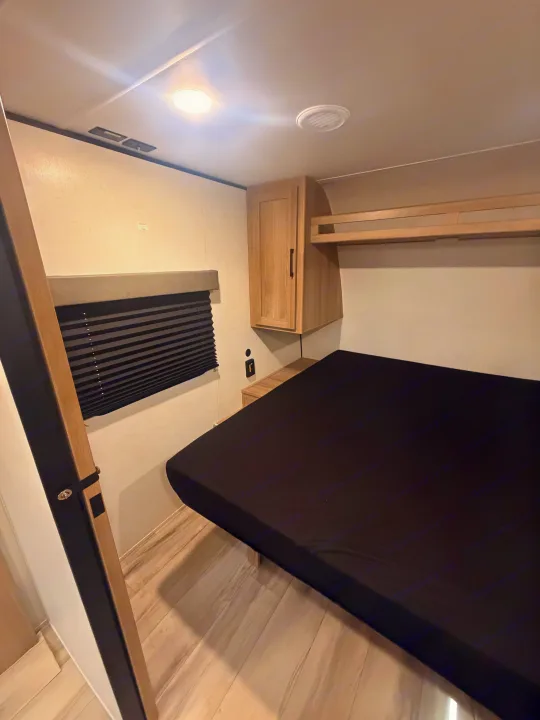 Modern RV rental bedroom with a compact bed, overhead storage, and window blind, perfect for travel.. Keystone Coleman 2026