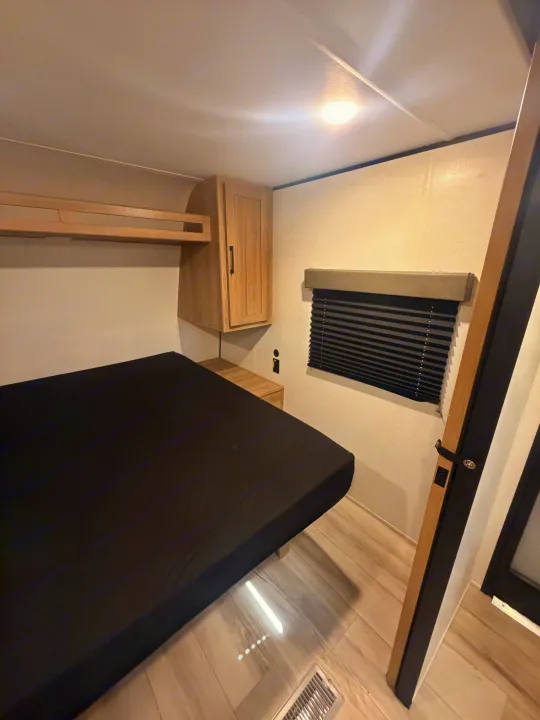 Compact RV rental bedroom featuring a black bed, light wood overhead cabinets, and a window with dark blinds.. Keystone Coleman 2026