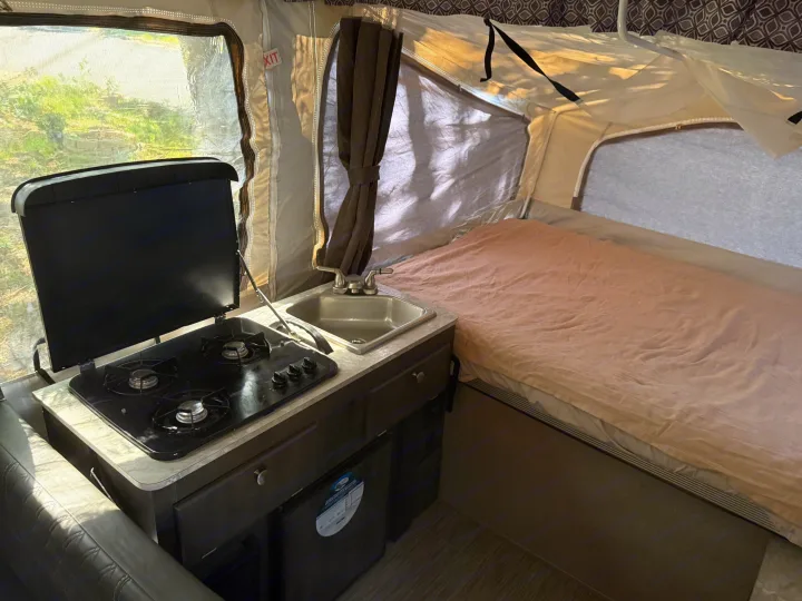 RV rental pop-up camper interior: kitchen with stove & sink, cozy bed with pink sheet, bright window view.. Forest River Flagstaff 2020