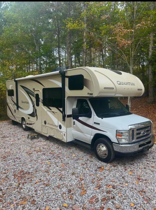 This beige Thor Quantum rv rental, a motorhome, is parked on a gravel campsite surrounded by trees.. Thor Quantum 2017
