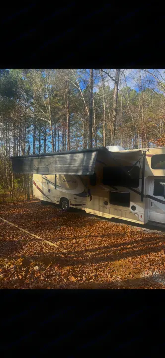 An RV rental, a Quantum RV, parked on a leaf-covered campsite with its awning extended, surrounded by tall trees.. Thor Quantum 2017