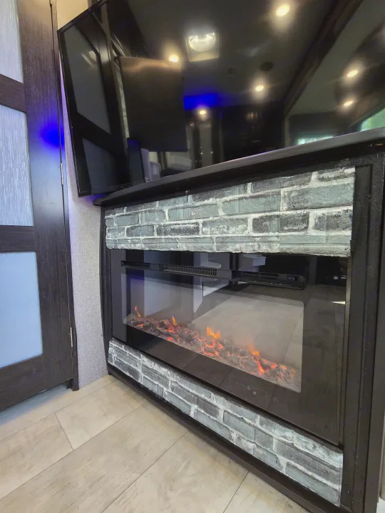 Modern RV rental interior with a glowing electric fireplace, overhead TV, and blue accent lighting, framed by faux brick.. Grand Design Momentum 2019