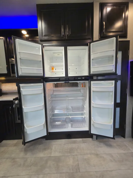 Spacious RV rental kitchen with a large, open multi-door refrigerator/freezer unit, showing empty shelves and storage for travel.. Grand Design Momentum 2019