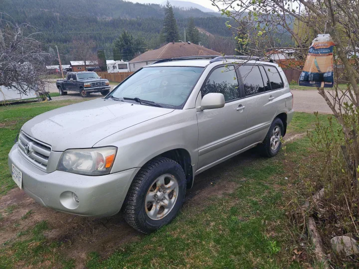 Rain-kissed silver SUV parked on grass next to a house with a truck and an RV in the background, possibly from an RV rental.. Toyota Highlander 2004