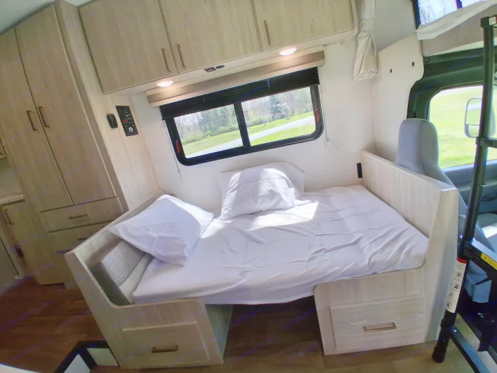 RV rental interior features a cozy bed with white linens, built-in storage, and a large window overlooking scenery.. East to West Entrada 2026