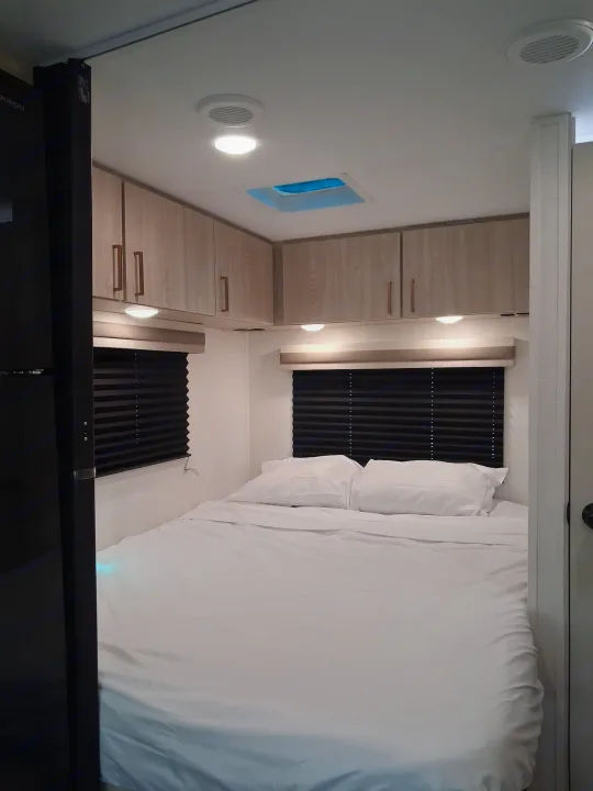 RV rental bedroom: cozy bed with white linens, black blinds, overhead cabinets, and bright ceiling & under-cabinet lighting.. East to West Entrada 2026