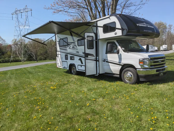 A white Forest River East West RV rental with its awning extended, parked on a sunny, dandelion-dotted lawn.. East to West Entrada 2026