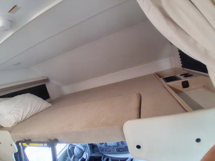 Overhead bunk in an rv rental, featuring a pillow, compact sink/stove area, and vehicle dashboard below.. East to West Entrada 2027