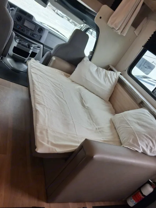 Interior view of an rv rental showing a comfortable bed with pillows and white sheets, with the vehicle's dashboard ahead.. East to West Entrada 2027