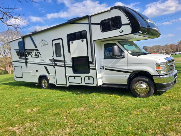 A white EastWest Entrada Class C RV rental, with black accents and mountain graphics, parked on grassy ground under a blue sky.. East to West Entrada 2027