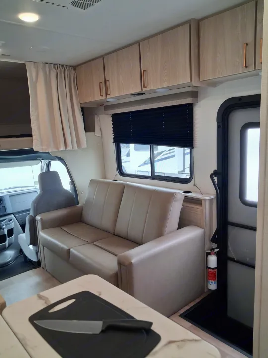 This rv rental interior features a comfortable sofa, overhead cabinets, a kitchen counter, and safety gear near the entry door.. East to West Entrada 2027