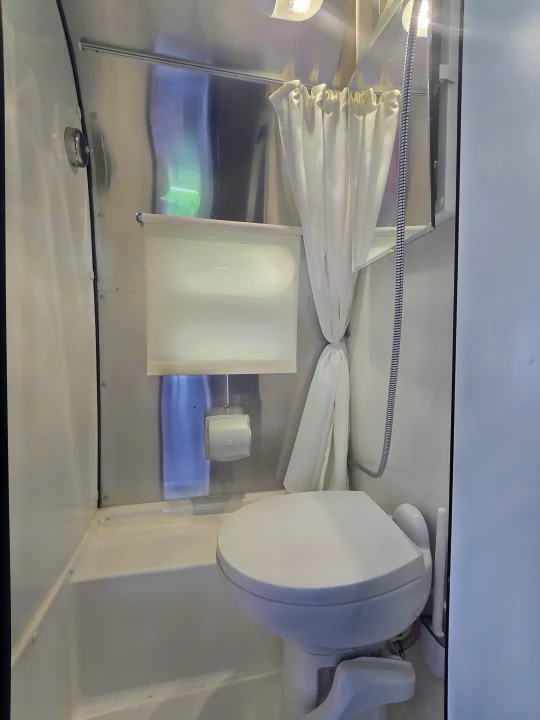 Compact RV rental bathroom featuring a toilet, shower with curtain, and mirrored cabinet on metallic walls, optimizing space.. Airstream Caraval 2021