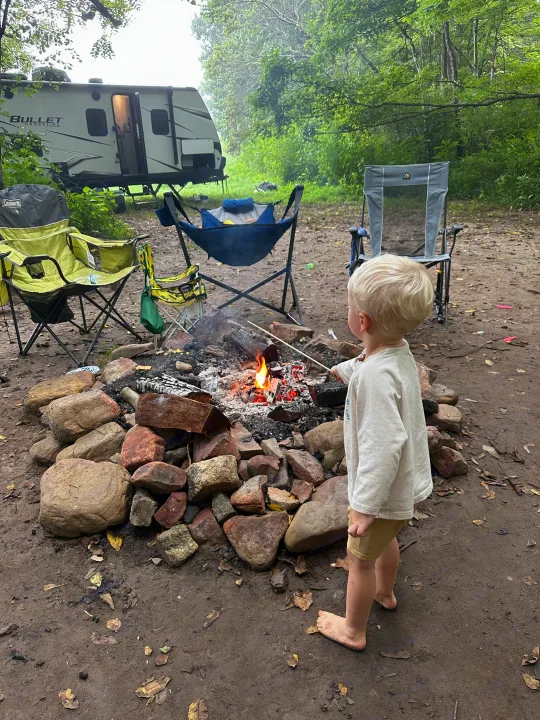 Young child pokes a glowing campfire at an RV rental site. A Bullet Crossfire travel trailer and camping chairs are visible.. Keystone RV Bullet Crossfire 2021