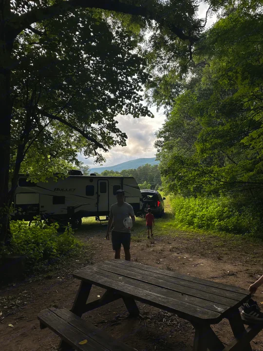 Family time at an RV rental campsite. A man and child stand by a travel trailer and picnic table, framed by trees and mountains.. Keystone RV Bullet Crossfire 2021