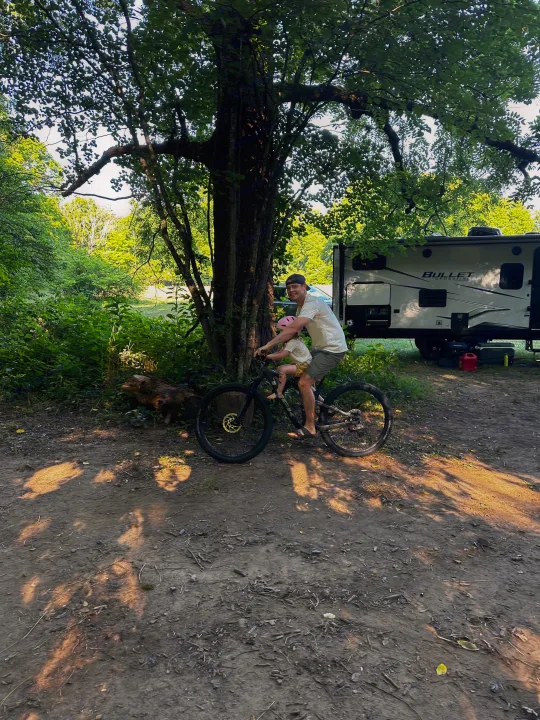 Dad and child enjoy a bike ride at their RV rental campsite, featuring a Bullet Crossfire travel trailer in the background.. Keystone RV Bullet Crossfire 2021