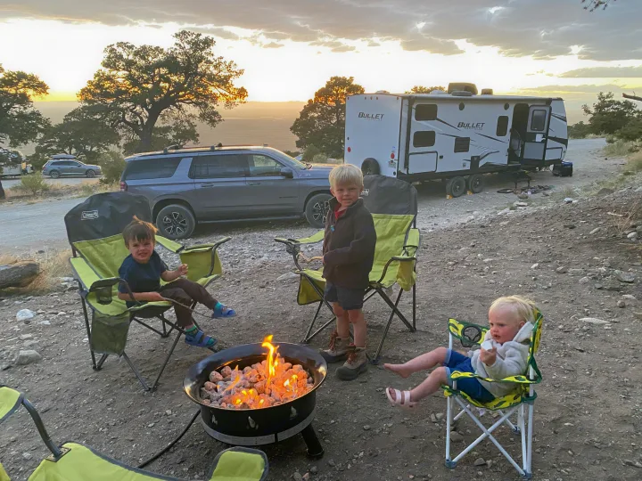 Young boys enjoy a warm campfire at their RV rental campsite, a travel trailer and SUV parked nearby at sunset.. Keystone RV Bullet Crossfire 2021