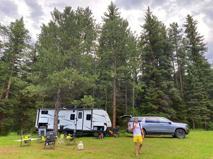 Man at his rv rental campsite with a white travel trailer, grey SUV, chairs, and bikes under tall pines.. Keystone RV Bullet Crossfire 2021