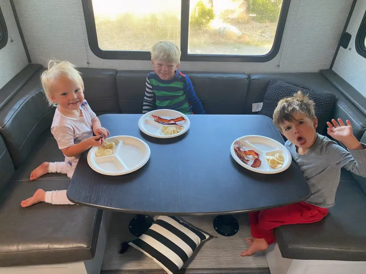 Three young children eat bacon & bananas at an RV rental dinette, smiling for a fun family breakfast while traveling.. Keystone RV Bullet Crossfire 2021