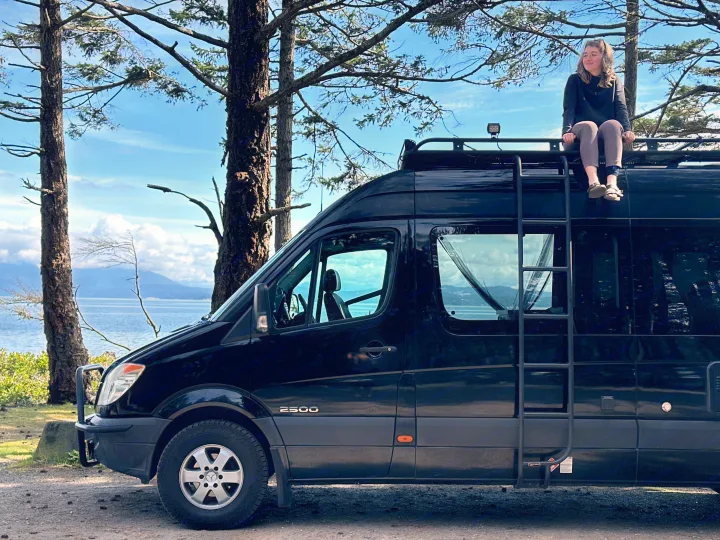 A woman sits atop a black RV rental van, enjoying breathtaking ocean and mountain views from the roof rack.. Mercedes Sprinter 2007