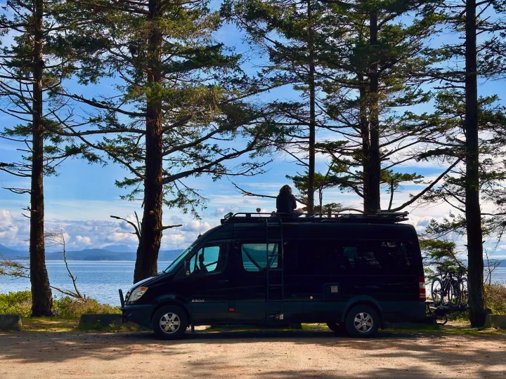 Black RV rental van by the ocean, with a person on the roof and bikes on the back, surrounded by tall trees.. Mercedes Sprinter 2007