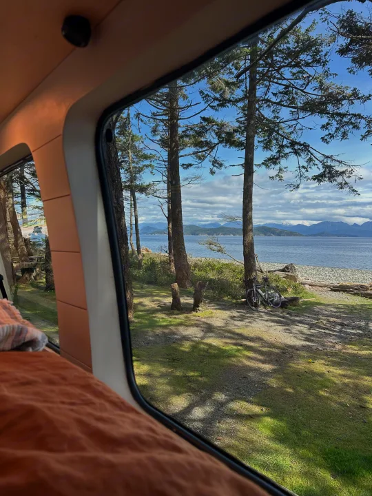 A tranquil scene from an RV rental: calm ocean, distant snowy peaks, and tall trees under a partly cloudy sky. A bike awaits adventure.. Mercedes Sprinter 2007