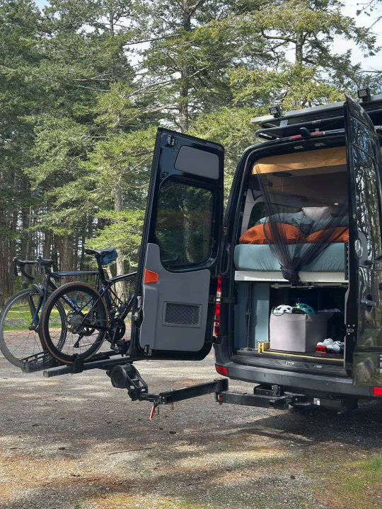 Black camper van RV rental, ready for adventure! Rear doors open to a cozy bed, bikes on rack, parked among tall forest trees.. Mercedes Sprinter 2007