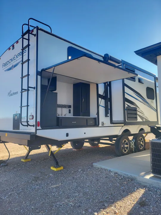 White Coachmen Freedom Express rv rental travel trailer with side open, showing an outdoor kitchen. Parked on gravel.. Coachmen Freedom Express liberty edition 326bhdsle 2023